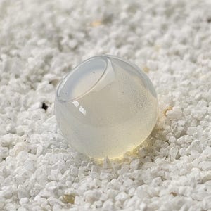 Pode incluir: Um objeto de vidro transparente e arredondado, com um interior oco, repousando sobre uma cama de pequenas pedras brancas. O vidro tem uma aparência ligeiramente leitosa, e a luz reflete-se na sua superfície lisa.