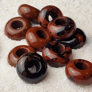 May include: A collection of brown and black donut-shaped beads with a hole in the center. The beads are made of a natural stone with a speckled pattern.