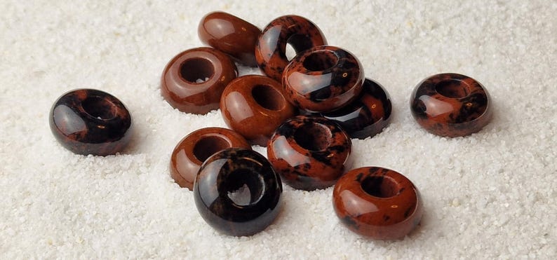 May include: A collection of brown and black donut-shaped beads with a hole in the center. The beads are made of a natural stone with a speckled pattern.