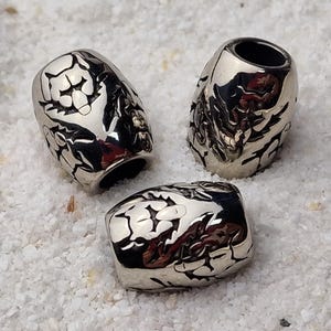 Könnte beinhalten: Drei silberfarbene Metallperlen mit detailliertem, strukturiertem Design. Die Perlen haben eine zylindrische Form mit einem hohlen Zentrum. Das Design scheint ein stilisiertes Muster zu sein. Die Perlen werden auf einer weißen, strukturierten Oberfläche präsentiert.