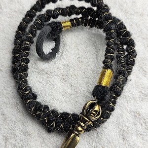 May include: A black and gold beaded necklace with a decorative bronze pendant. The necklace is made of black beads with gold accents and a bronze pendant with a pointed end. The necklace is displayed on a white surface.