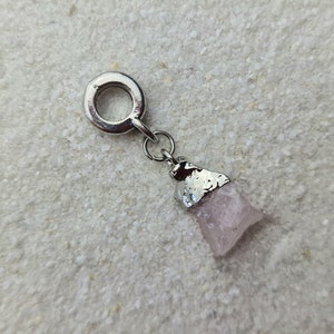 Op de afbeelding: Een zilveren bedel met een roze kristal eraan bevestigd. De bedel heeft een lus om aan een armband of ketting te bevestigen.