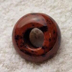 May include: A red and black donut-shaped bead with a hole in the center. The bead is made of polished stone and has a glossy finish.