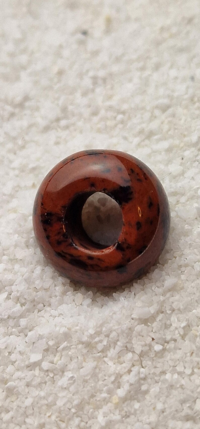 May include: A red and black donut-shaped bead with a hole in the center. The bead is made of polished stone and has a glossy finish.