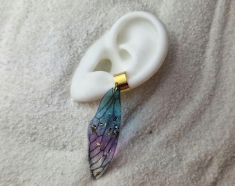 May include: A pair of iridescent blue and purple dragonfly wing earrings with gold accents. The earrings are hanging from a gold hoop.