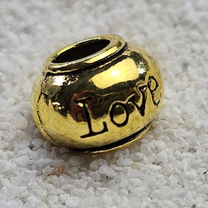 Op de afbeelding: Een goudkleurige kraal met het woord "Love" op het oppervlak gegraveerd. De kraal heeft een afgeronde vorm met een cilindrische bovenkant en ligt op een bed van witte kiezelstenen. De kraal is reflecterend.