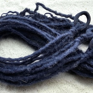 Op de afbeelding: Een bundel donkerblauwe dreadlocks met elastiekjes aan de uiteinden, liggend op een lichtgekleurd, getextureerd oppervlak. De dreadlocks zijn dik en hebben een enigszins vage textuur.