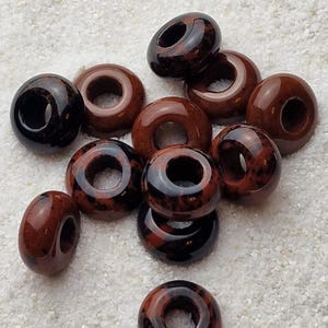 May include: A group of ten brown and black donut-shaped ceramic beads with a hole in the center. The beads are scattered on a white surface.