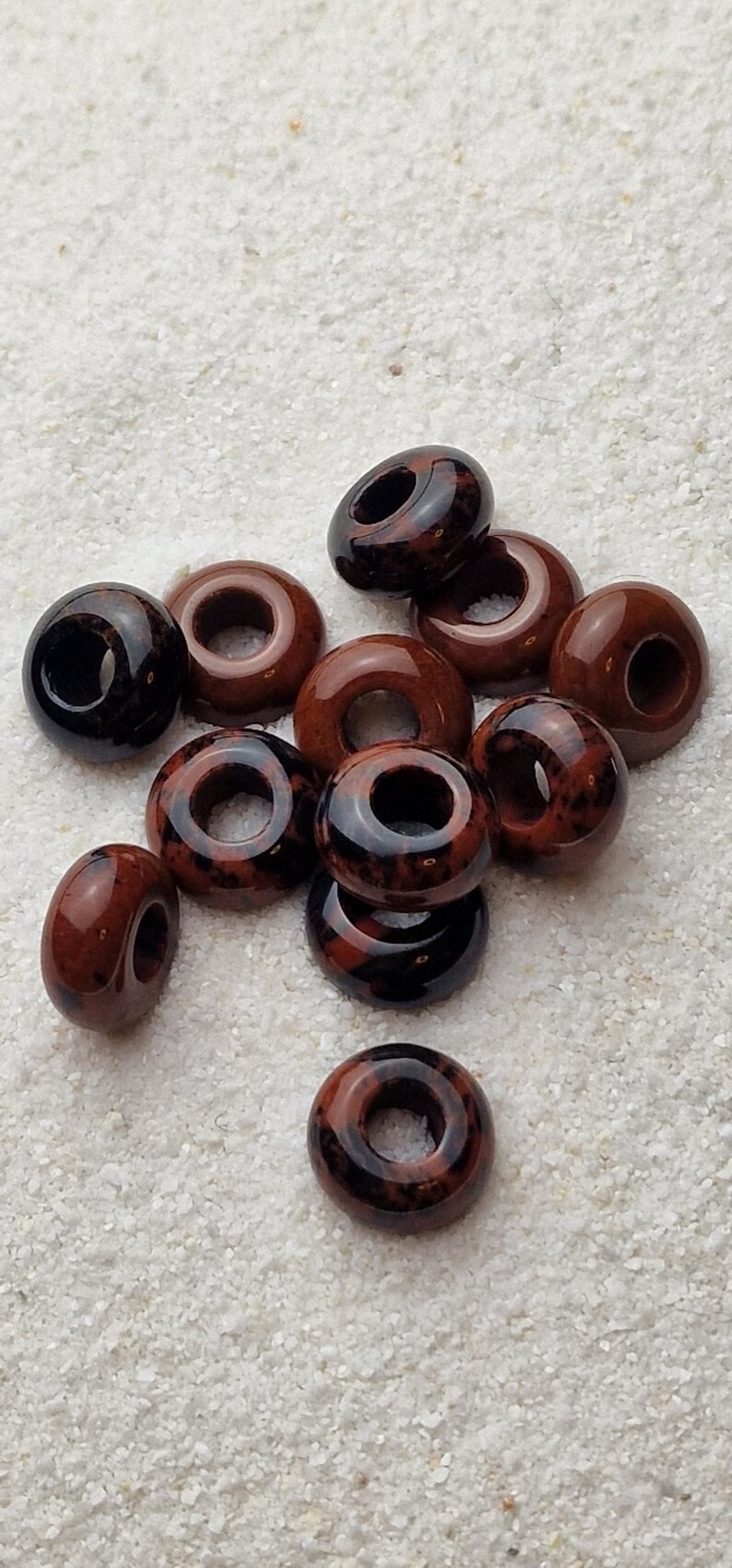 May include: A group of ten brown and black donut-shaped ceramic beads with a hole in the center. The beads are scattered on a white surface.