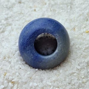May include: A blue ceramic bead with a hole in the center. The bead has a light blue glaze on one side and a darker blue glaze on the other side.