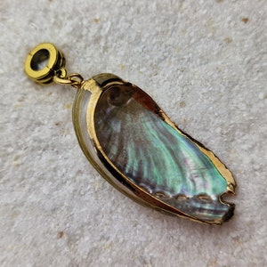 Puede incluir: Colgante de concha de abulón con iridiscencias azules, verdes y ribete dorado. La concha está encerrada en un marco de metal dorado y tiene un pequeño lazo en la parte superior para una cadena. El colgante se muestra sobre una superficie texturizada blanquecina.