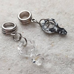 Op de afbeelding: Twee zilveren bedelarmbanden met een zilveren ketting die ze verbindt. Eén armband heeft een heldere kristallen bedel eraan bevestigd, en de andere armband heeft een zwart-witte bedel eraan bevestigd.