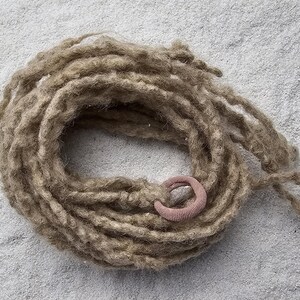 Op de afbeelding: Een opgerolde bundel lichtbruine dreadlocks met een kleine, roze haarband. De dreadlocks zijn gemaakt van een textuurrijk materiaal en zijn in een cirkelvorm gerangschikt. De achtergrond is een lichtgrijze, getextureerde ondergrond.