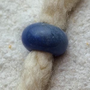 May include: A close-up of a blue bead on a white braided cord. The bead is smooth and round, and the cord is made of natural fibers.