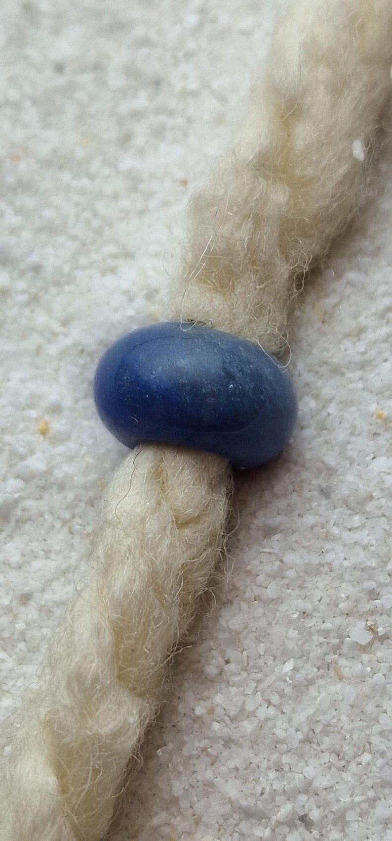 May include: A close-up of a blue bead on a white braided cord. The bead is smooth and round, and the cord is made of natural fibers.