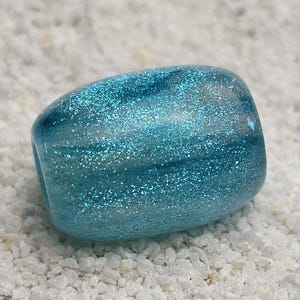 Könnte beinhalten: Eine blau-grüne, abgerundete rechteckige Perle mit glitzernden Akzenten. Die Perle hat eine leicht gebogene Form und liegt auf einem weißen, strukturierten Hintergrund. Die Farbe der Perle geht von einem dunkleren Blau in ein helleres, schimmerndes Türkis über.