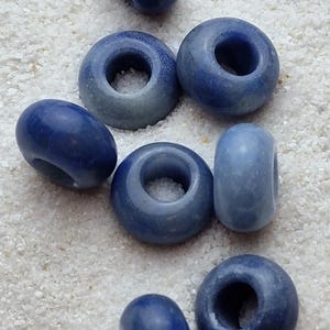 May include: A collection of eight blue ceramic beads with a hole in the center. The beads are various shades of blue and are arranged in a cluster on a white surface.