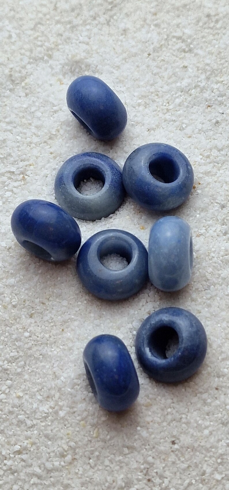 May include: A collection of eight blue ceramic beads with a hole in the center. The beads are various shades of blue and are arranged in a cluster on a white surface.
