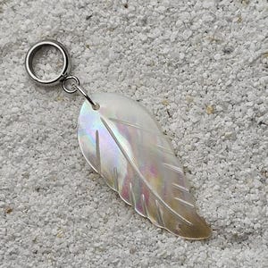 Könnte beinhalten: Ein perlmuttfarbener, blattförmiger Anhänger mit schillernden Farbtönen in Rosa, Lila und Gold, befestigt an einem silberfarbenen Ring. Der Anhänger liegt auf einer strukturierten, hellen Oberfläche.
