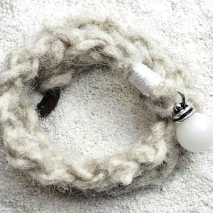 Könnte beinhalten: Ein geflochtenes, hellgraues Armband mit einem weißen, runden Anhänger und silberfarbenen Akzenten. Das Armband besteht aus dickem, strukturiertem Garn und hat einen dunkelbraunen Verschluss.