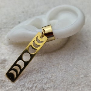 May include: Gold ear cuff with a dangling charm featuring a crescent moon design. The charm is a long, thin rectangle with eight different phases of the moon cut out.