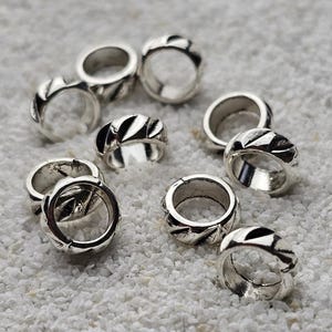 May include: A collection of silver rings with a textured design. The rings are round with a band featuring a series of diagonal cuts. The rings are scattered on a white, textured surface.