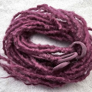 Op de afbeelding: Een bundel dieppaarse dreadlocks, opgerold en vastgezet met een bijpassende scrunchie. De dreadlocks hebben een getextureerd, handgemaakt uiterlijk en zijn gerangschikt op een lichtgrijze ondergrond.