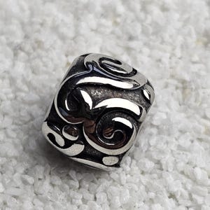 Pode incluir: Uma conta prateada com um design ornamental em espiral. A conta tem uma forma quadrada com bordas arredondadas e está sobre um fundo branco texturizado. O design apresenta uma série de linhas curvas interconectadas.