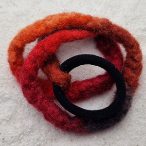 Op de afbeelding: Een close-up van een haarband gemaakt van vilten garen in tinten oranje, rood en zwart. De haarband heeft de vorm van een cirkel en ligt op een wit oppervlak.