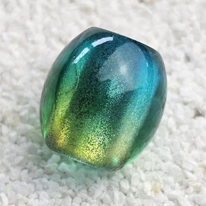 Könnte beinhalten: Eine abgerundete, irisierende Glasperle mit einem Farbverlauf aus Grün, Blau und Gelb. Die Perle ist glatt und reflektierend und liegt auf einem Bett aus kleinen, weißen Steinen. Der Gesamteffekt ist einer von Tiefe und Leuchtkraft.