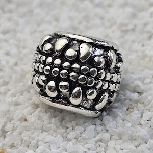 May include: A silver bead with a textured surface. The bead is square-shaped with a pattern of raised, irregular shapes and small circles. The bead is resting on a bed of small, white pebbles.