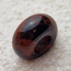 May include: A dark red and black gemstone bead with a hole drilled through the center. The bead is smooth and polished.