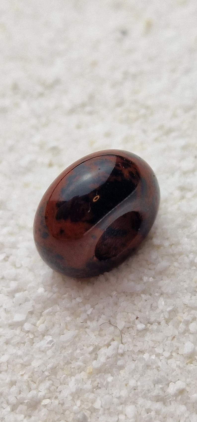May include: A dark red and black gemstone bead with a hole drilled through the center. The bead is smooth and polished.