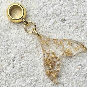 Puede incluir: Encanto dorado con una cola de sirena de resina transparente con purpurina dorada incrustada. El encanto está unido a un anillo dorado.