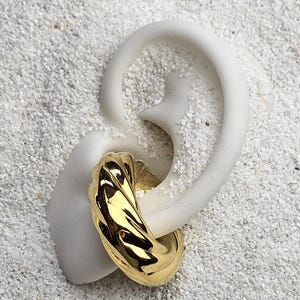 May include: A gold-colored, textured hoop earring is displayed on a white ear-shaped form. The earring has a ridged design and reflects light. The background is a textured, white surface, possibly sand.