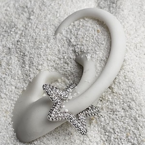 May include: A silver star-shaped earring with a textured, sparkling surface. The earring is displayed on a white, curved stand, set against a backdrop of small, white pebbles. The earring's design is contemporary and striking.