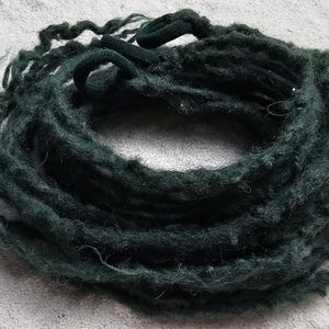 Könnte beinhalten: Ein Bündel dunkelgrüner Dreadlocks, eng gewickelt, liegt auf einer hellgrauen Oberfläche. Die Dreadlocks scheinen handgefertigt zu sein und haben ein leicht strukturiertes Aussehen. Der Gesamteindruck ist natürlich und bohem.