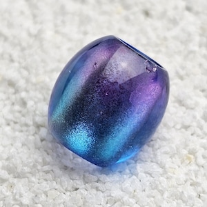 Könnte beinhalten: Eine handgefertigte, irisierende Glasperle mit einem Farbverlauf aus Blau, Lila und Türkis. Die ovale Perle liegt auf einem Bett aus kleinen, weißen Steinen. Die Oberfläche der Perle reflektiert das Licht und erzeugt einen Schimmereffekt.