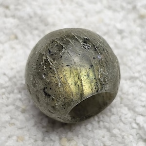 Puede incluir: Una cuenta esférica de aspecto metálico, con un agujero en el centro. La cuenta tiene un aspecto moteado con tonos de gris, plata y toques de amarillo. Probablemente hecha de una piedra natural o mineral.