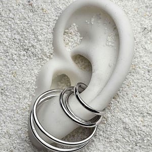 Può includere: Orecchini a cerchio color argento esposti su un modello di orecchio bianco su uno sfondo strutturato di colore chiaro. Gli orecchini hanno un design a strati, con tre cerchi collegati di varie dimensioni.