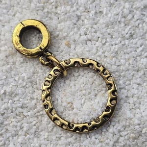 Könnte beinhalten: Ein goldfarbener Metallanhänger mit kreisförmigem Design. Der Anhänger hat oben eine kleine, runde Schlaufe und unten einen größeren, gemusterten Kreis. Das Muster besteht aus kleinen, gleichmäßig verteilten Vertiefungen.