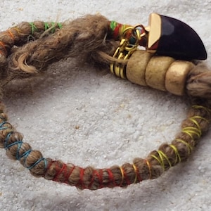 Könnte beinhalten: Ein brauner und beiger Dreadlock mit einem bunten Band darum gewickelt. Eine Holzperle mit einem goldenen Metallanhänger ist an dem Band befestigt.