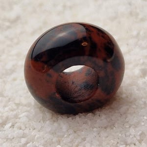 May include: A dark red and black donut-shaped bead with a hole in the center. The bead is made of natural stone and has a unique marbled pattern.