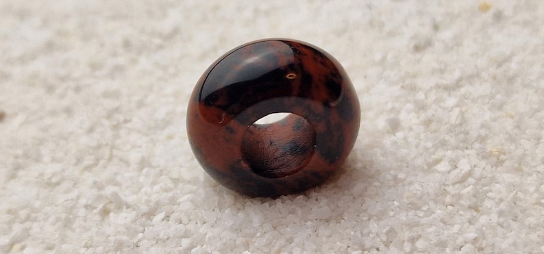 May include: A dark red and black donut-shaped bead with a hole in the center. The bead is made of natural stone and has a unique marbled pattern.