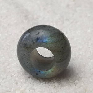 Könnte beinhalten: Ein dunkelgrauer donutförmiger Perle aus Labradorit mit schillernden blauen und grünen Farbtönen.