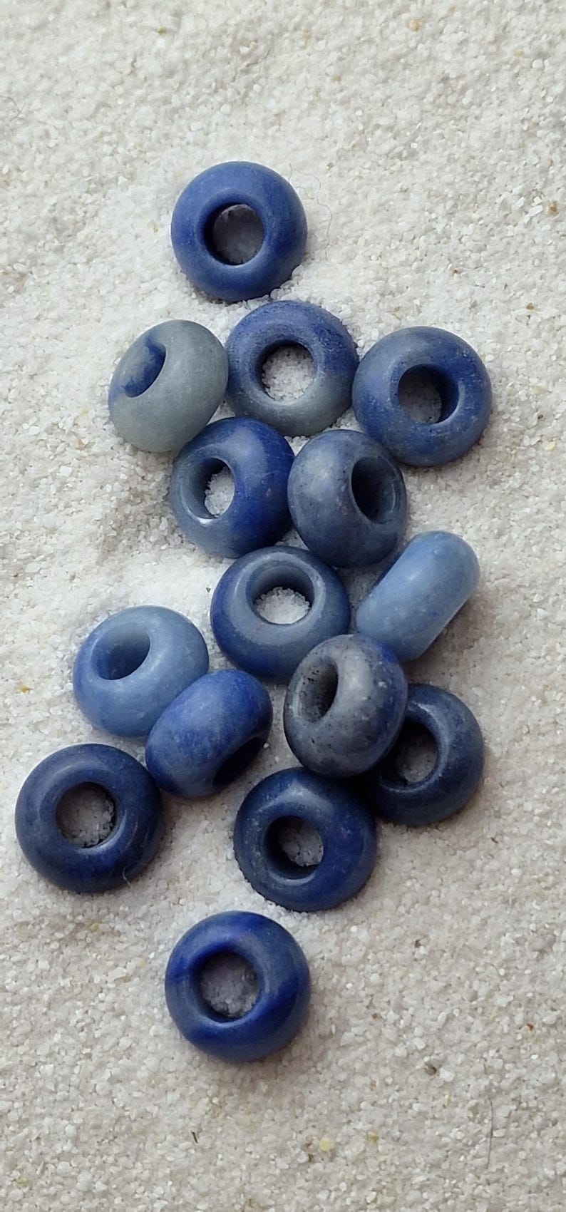 May include: A collection of blue and light blue donut-shaped beads with a hole in the center. The beads are arranged in a pile on a white surface.