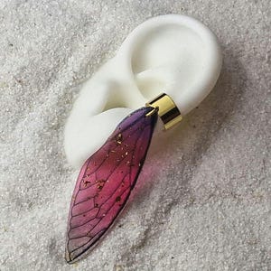 Pode incluir: Um brinco de ouro com um charme em forma de asa iridescente roxo e rosa. O charme tem manchas douradas por toda parte.