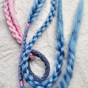Könnte beinhalten: Ein Haaraccessoire mit synthetischen Faserzöpfen in Pastellrosa und Hellblau. Die Zöpfe sind an einem dunkelgrauen Haargummi befestigt, wobei Abschnitte des blauen Zopfes mit rosa Faden umwickelt sind, um die Haarsträhnen zusammenzuhalten.