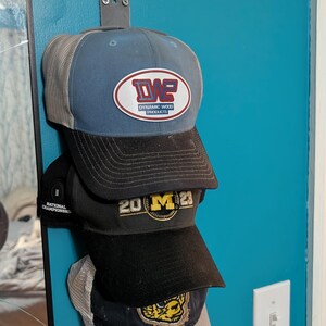 May include: A stack of baseball caps in various colors, including blue, black, and gray, hanging on a wall. The top cap has a logo that reads "DWE Dynamic Wood Products." Other caps display "20 M 23" and a tiger logo.