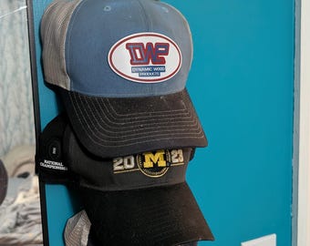 May include: A stack of baseball caps in various colors, including blue, black, and gray, hanging on a wall. The top cap has a logo that reads "DWE Dynamic Wood Products." Other caps display "20 M 23" and a tiger logo.